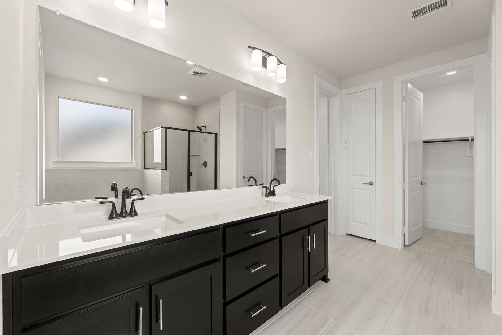 A modern, minimalist bathroom with a large vanity, double sinks, and a large mirror on the wall. The room has a clean, neutral color palette with white walls and dark cabinets.