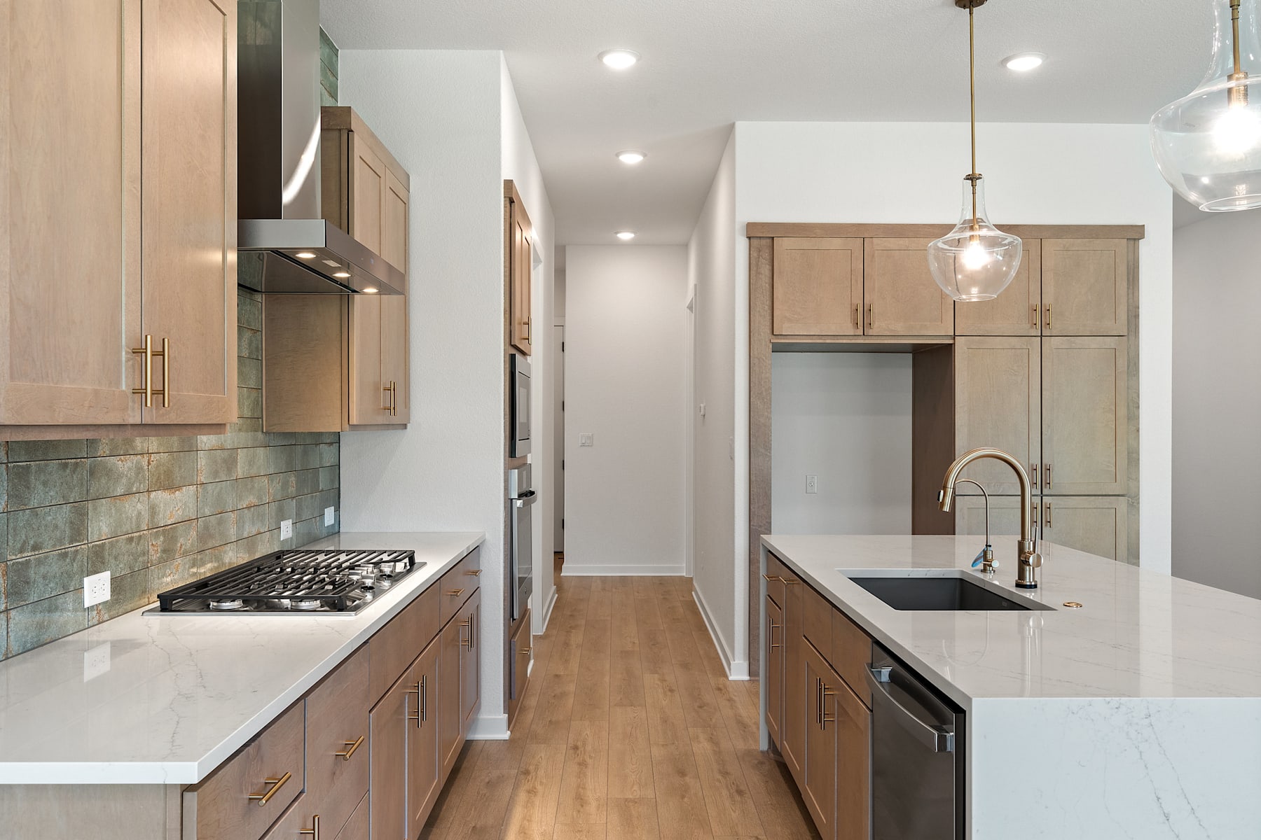 A modern, well-lit kitchen with light-colored wood cabinets, a tile backsplash, and stainless steel appliances, leading into a hallway with additional storage space.