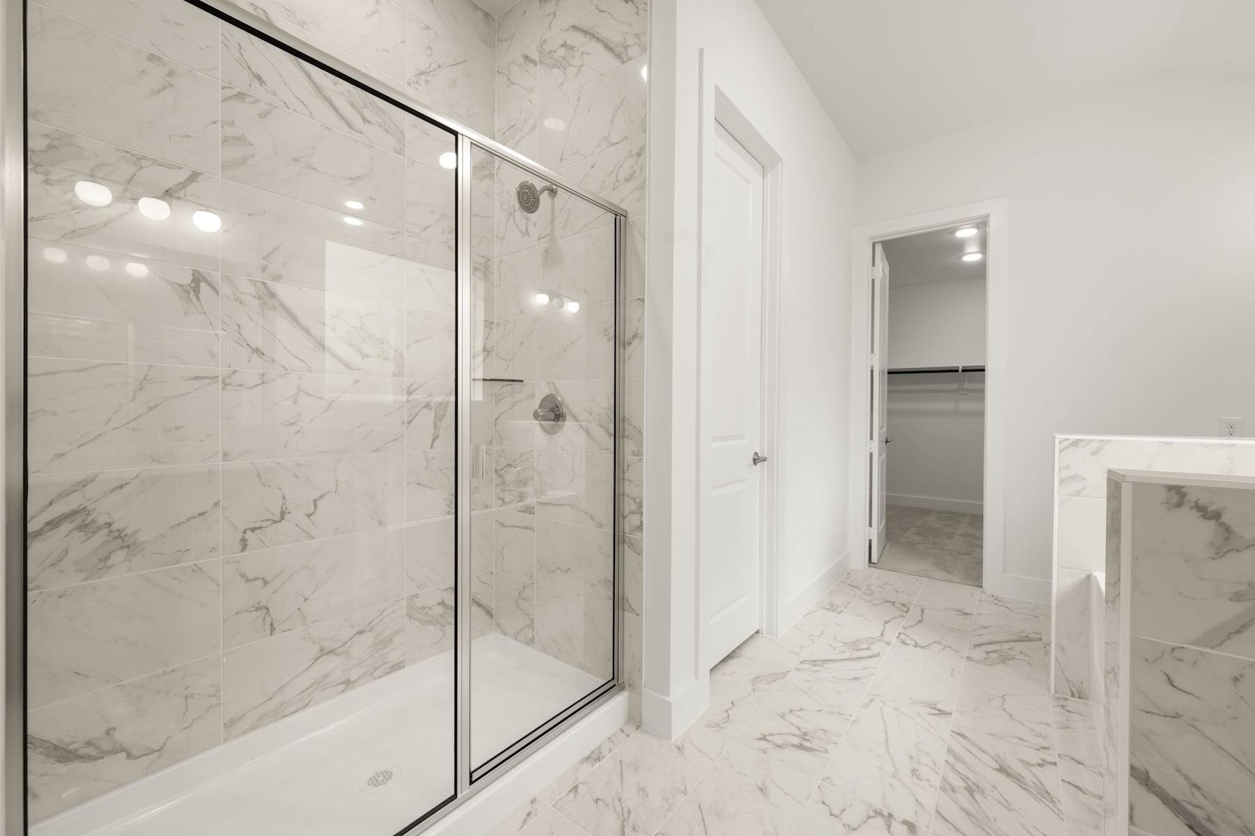 A modern, minimalist bathroom with sleek, white marble walls and a glass shower enclosure, leading to a hallway in the background.