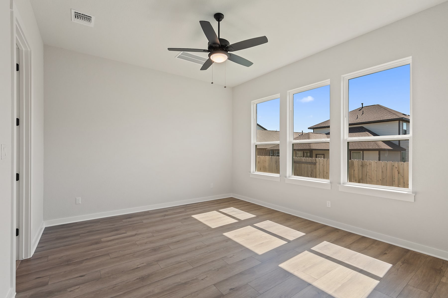 A bright and airy room with a ceiling fan, hardwood floors, and large windows overlooking a residential neighborhood.