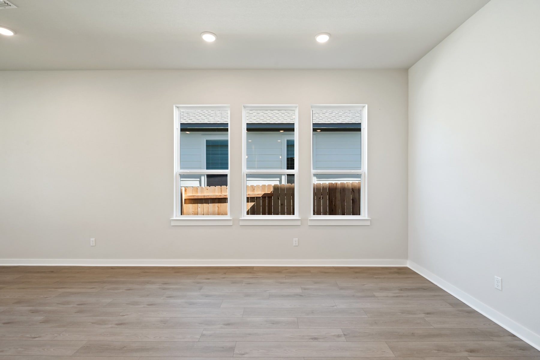 A spacious, well-lit room with white walls, wooden flooring, and three large windows overlooking an outdoor area.
