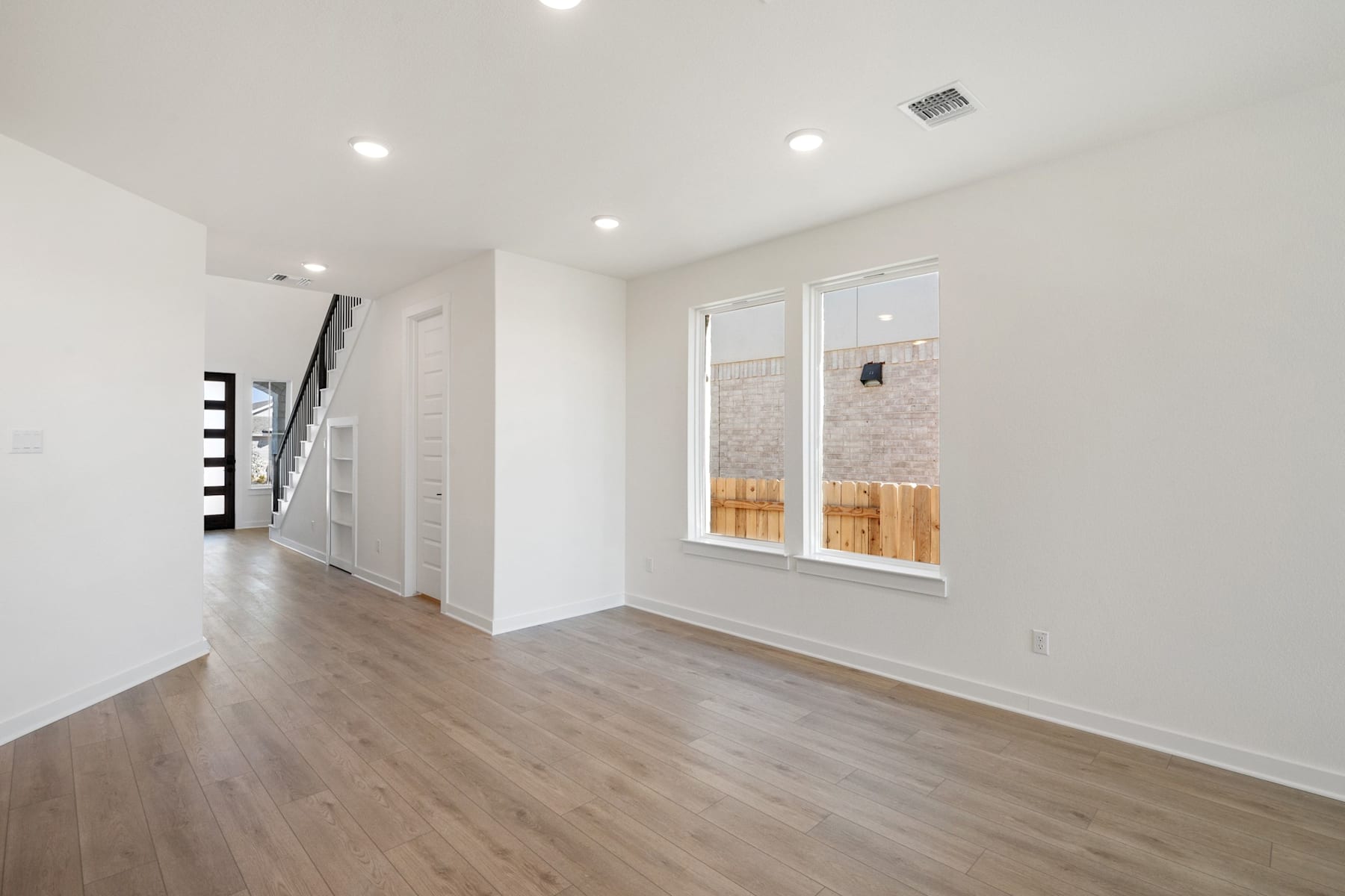 A spacious, well-lit room with white walls, hardwood floors, and recessed lighting. The room appears to be a living or dining area, with a window providing natural light and a view of the exterior.