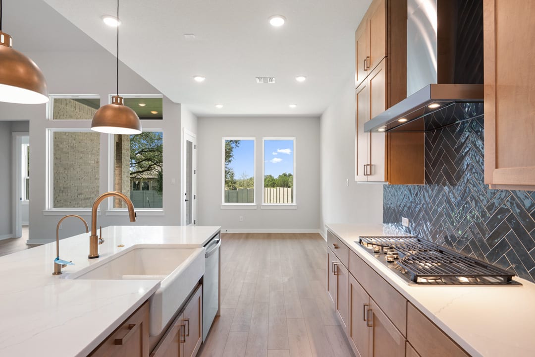 A modern, well-lit kitchen with a sleek design, featuring a sink, stove, and cabinets, with a view of the outdoors through a window in the background.