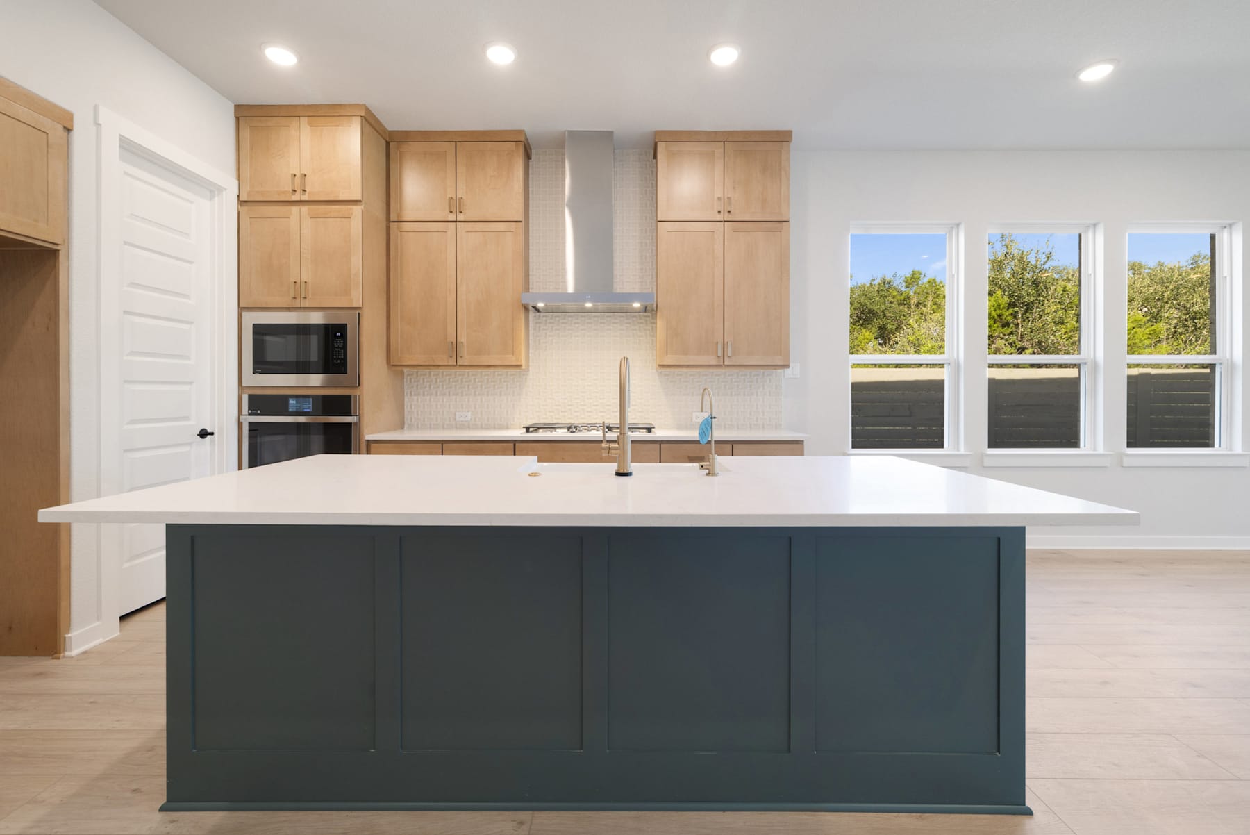 A modern, open-concept kitchen with light wood cabinets, a large white island, and a view of the outdoors through large windows.