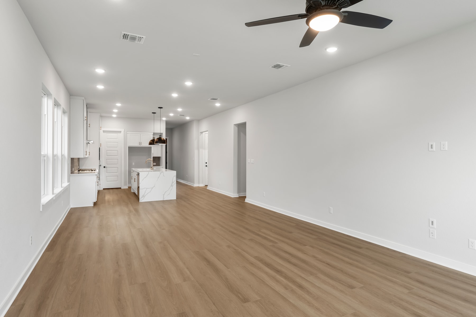A spacious, well-lit room with a hardwood floor, white walls, and a ceiling fan, leading into a kitchen area with white cabinets and appliances.