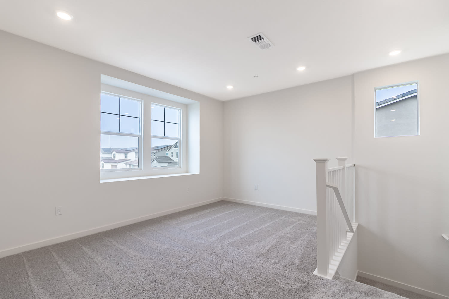 A bright, spacious room with white walls, a large window, and a carpeted floor. The room appears to be a bedroom or living space with a staircase visible in the background.