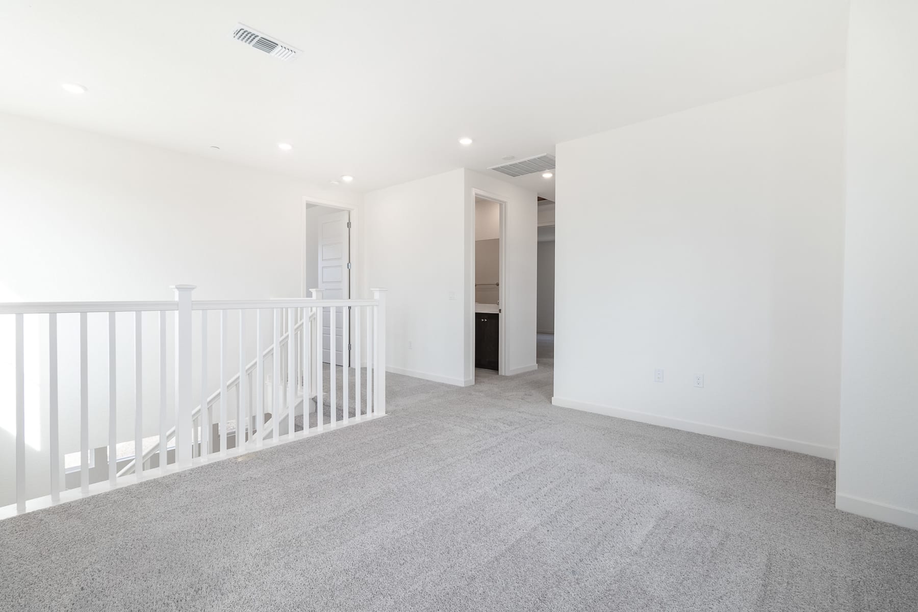 A bright, open hallway with a white railing leading to a doorway in the background, and a carpeted floor in the foreground.
