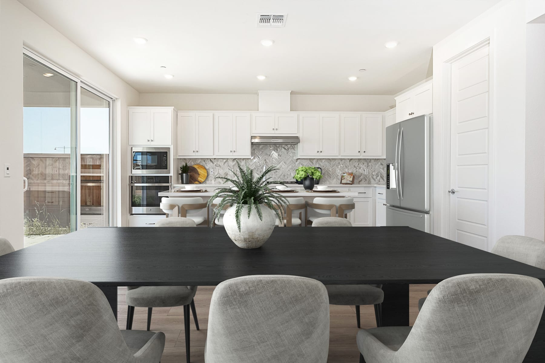 A modern, open-concept kitchen and dining area with white cabinets, stainless steel appliances, and a large black dining table surrounded by gray upholstered chairs.