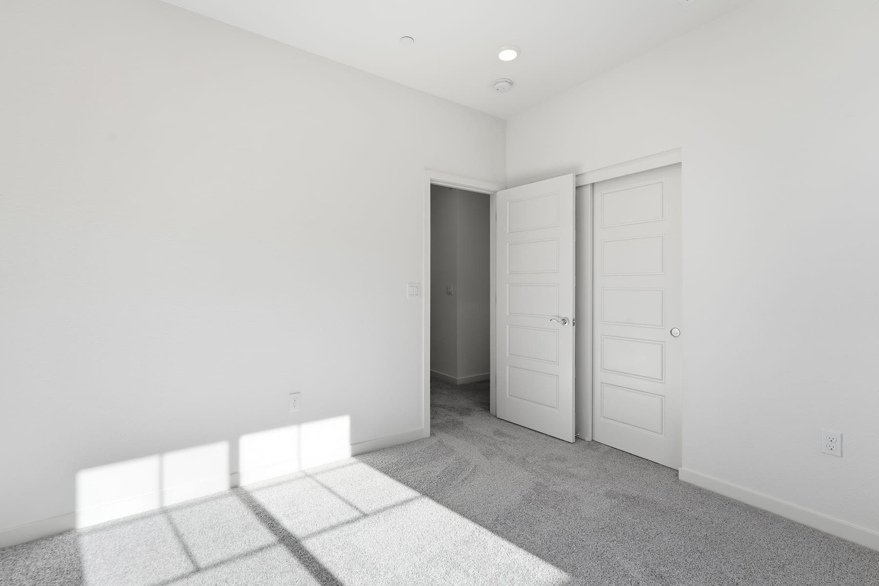 A simple, minimalist room with white walls, a closed door, and sunlight streaming in through a window, casting shadows on the gray carpeted floor.