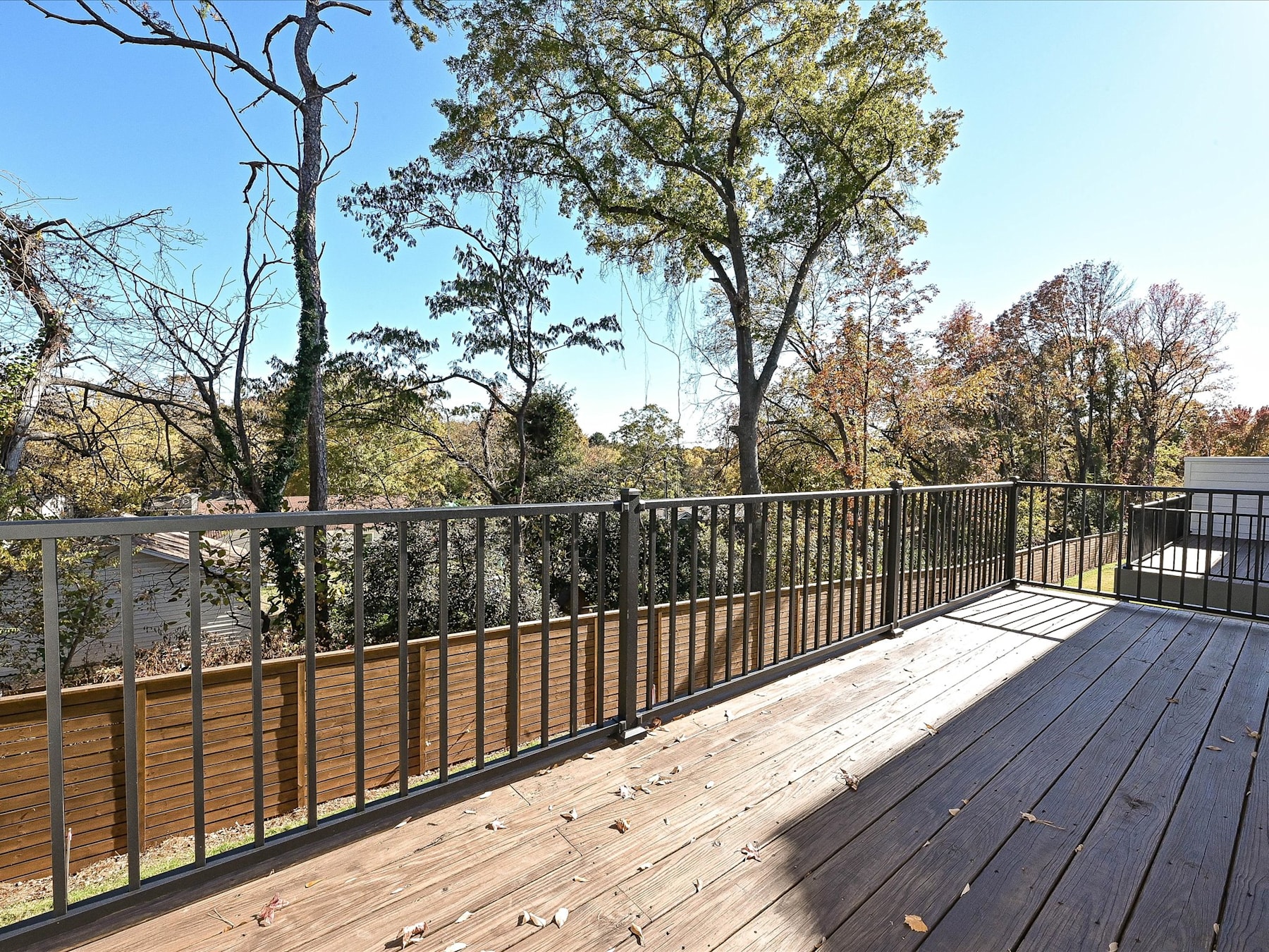 A wooden deck with a metal railing overlooks a wooded area with trees in various stages of foliage, set against a clear blue sky.