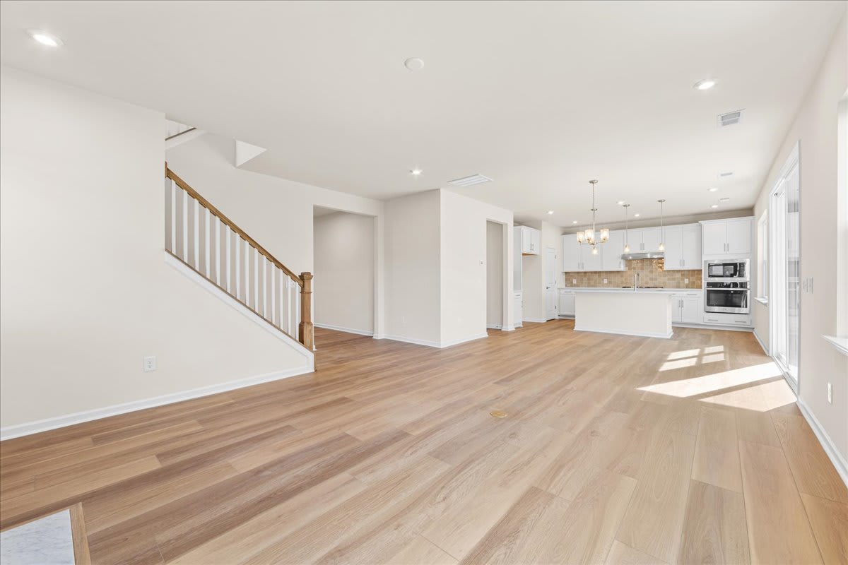 A spacious and bright open-concept living area with hardwood floors, a staircase, and a kitchen visible in the background.