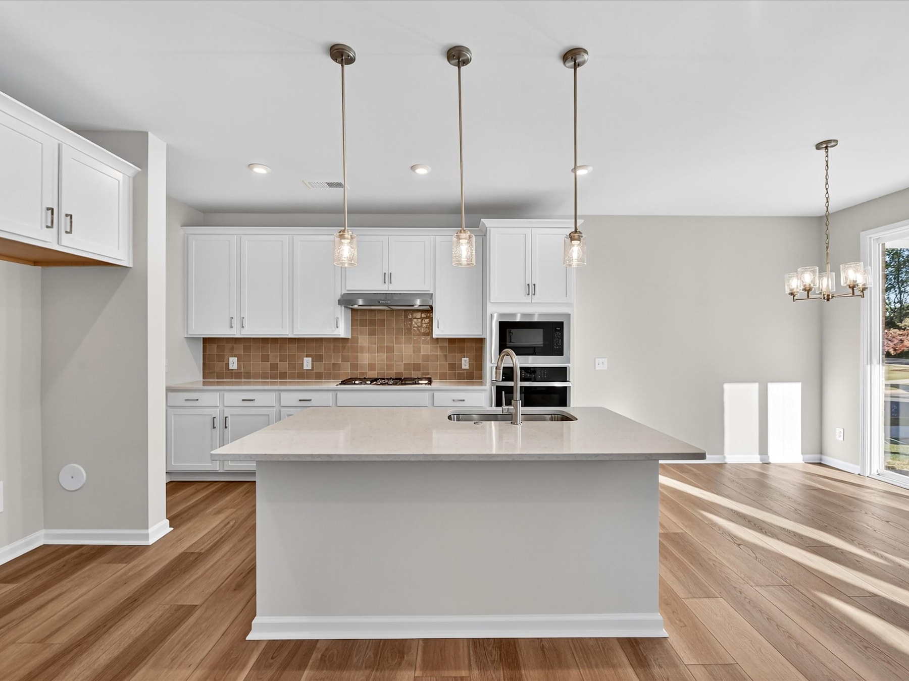 A modern, open-concept kitchen with white cabinets, a large island, and pendant lighting fixtures, set against a hardwood floor and neutral-toned walls.