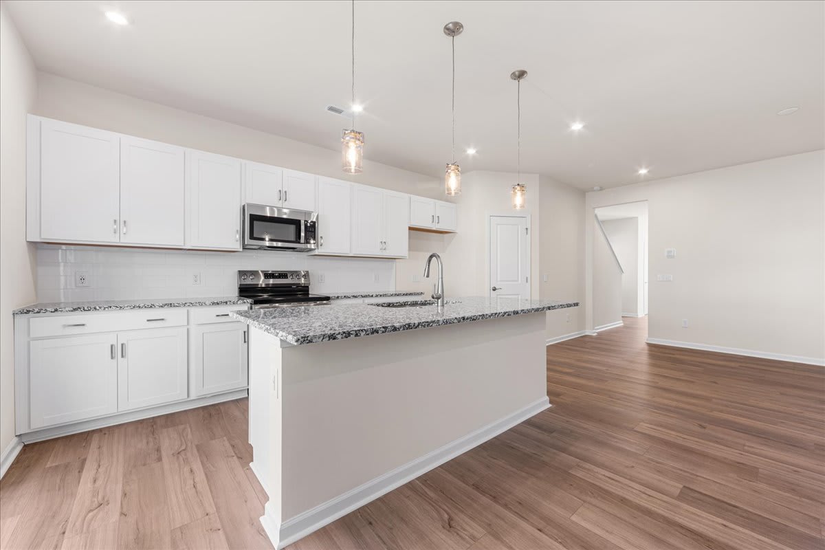 A modern, open-concept kitchen with white cabinets, granite countertops, and hardwood floors, illuminated by pendant lights.