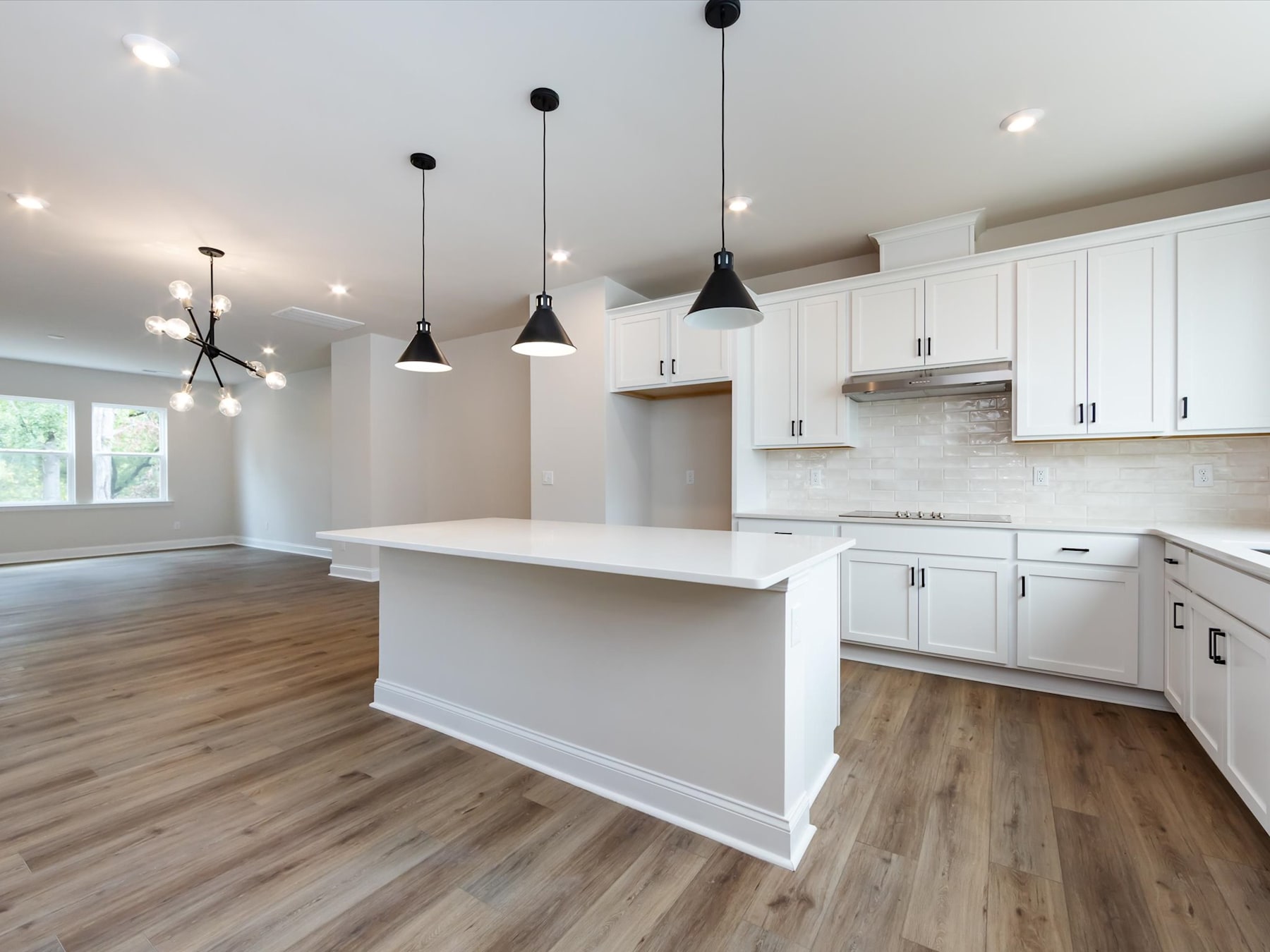 A modern, open-concept kitchen with white cabinets, a large island, and pendant lighting fixtures, set against a hardwood floor.