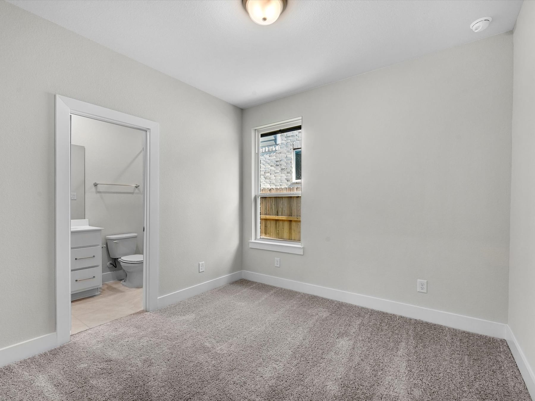 A simple, minimalist bedroom with a window, a door leading to an adjacent room, and a light fixture on the ceiling.