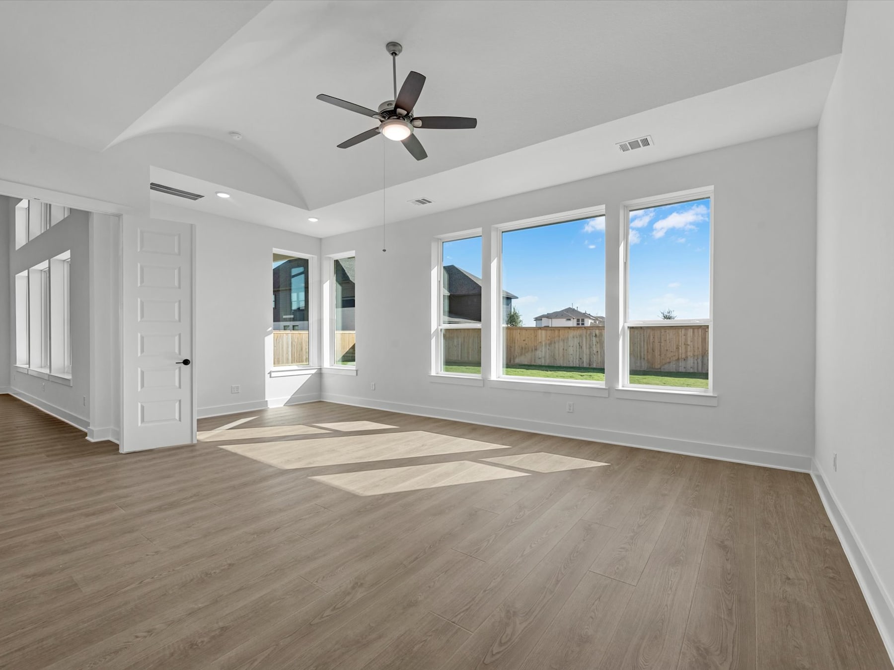 A spacious, well-lit room with a ceiling fan, hardwood floors, and large windows overlooking a fenced backyard.