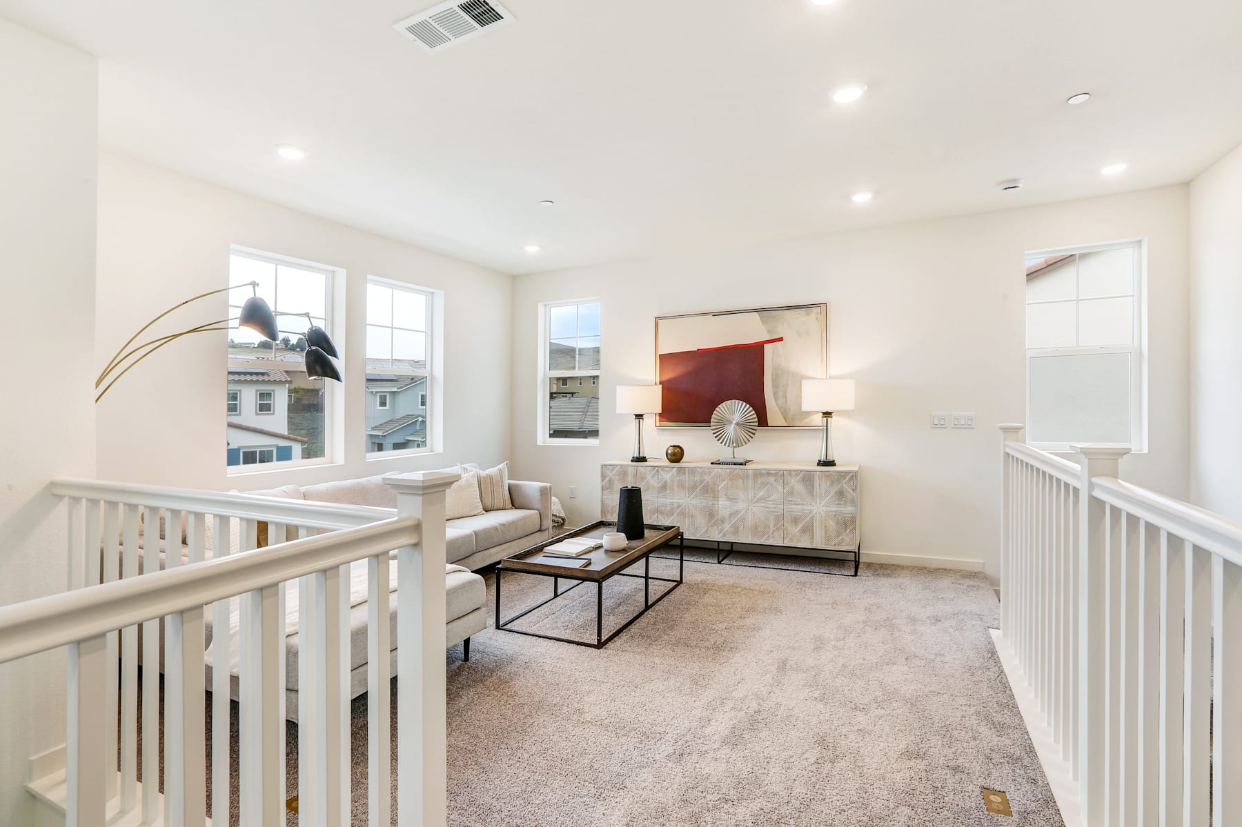 A bright and airy living room with a white railing and a cozy seating area in the foreground, and a decorative cabinet and artwork on the wall in the background.