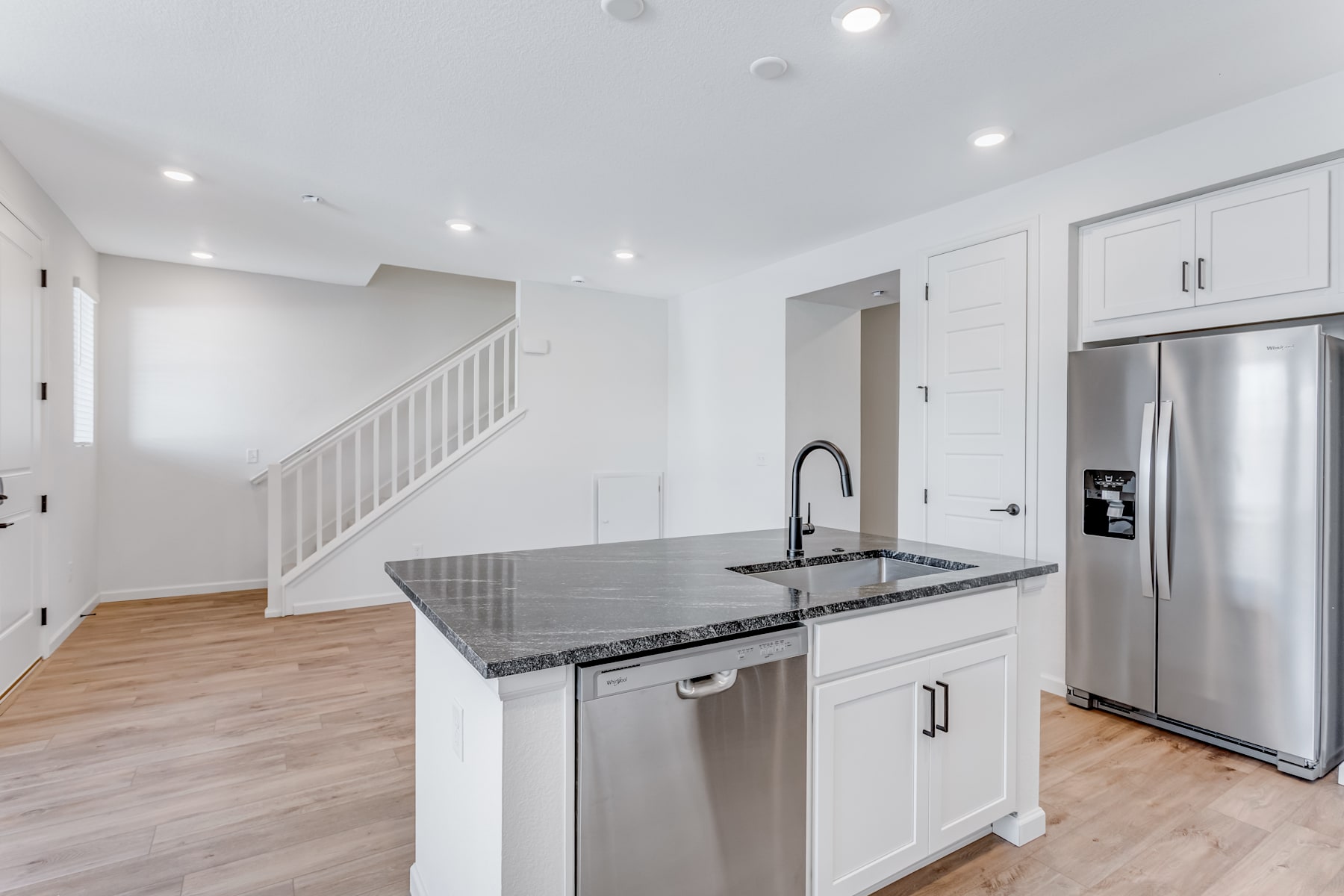 A modern, open-concept kitchen with white cabinets, stainless steel appliances, and a granite countertop, leading into a living area with hardwood floors and a staircase.