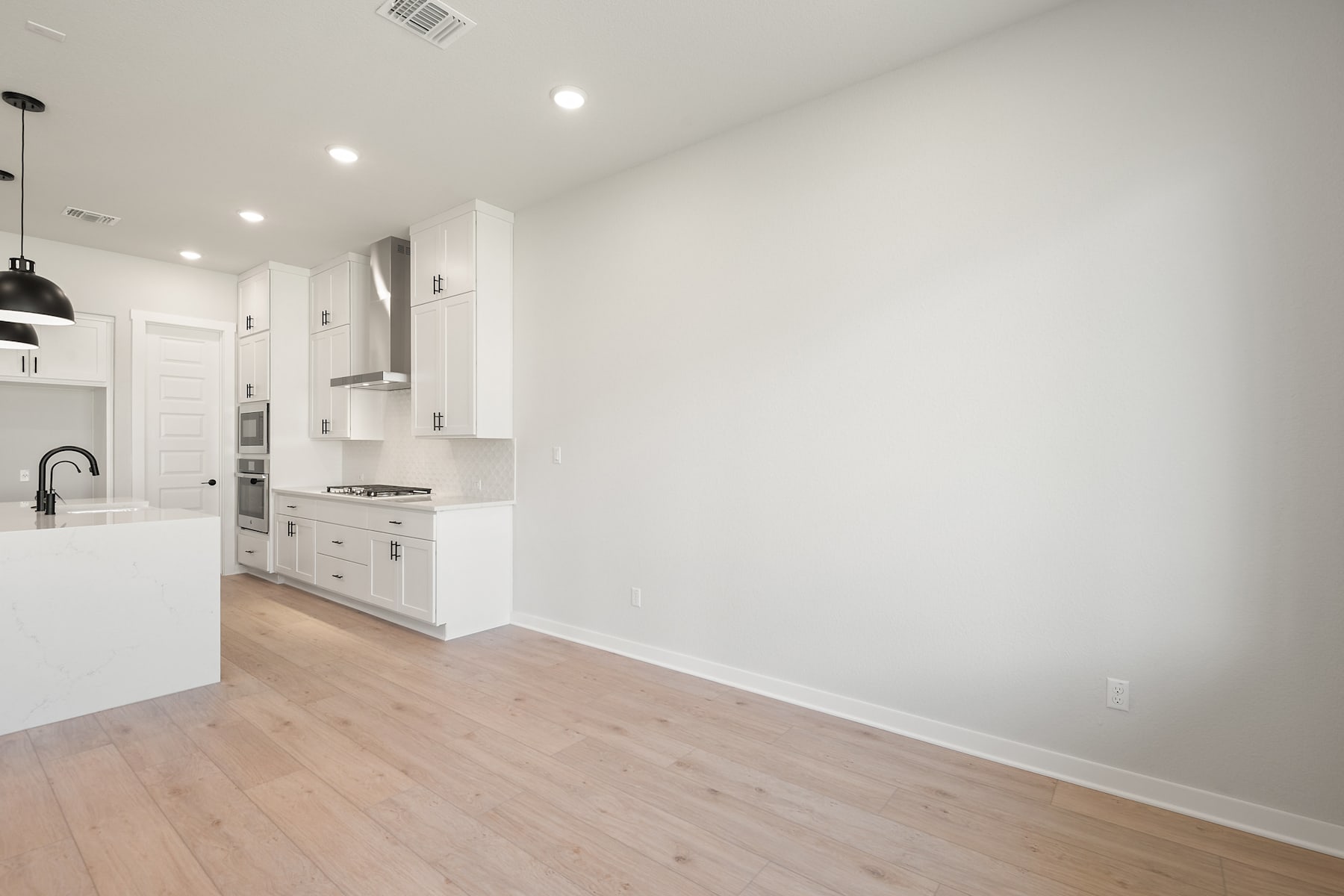 A modern, minimalist kitchen with white cabinets, stainless steel appliances, and hardwood floors, leading into an open living space with a blank white wall.