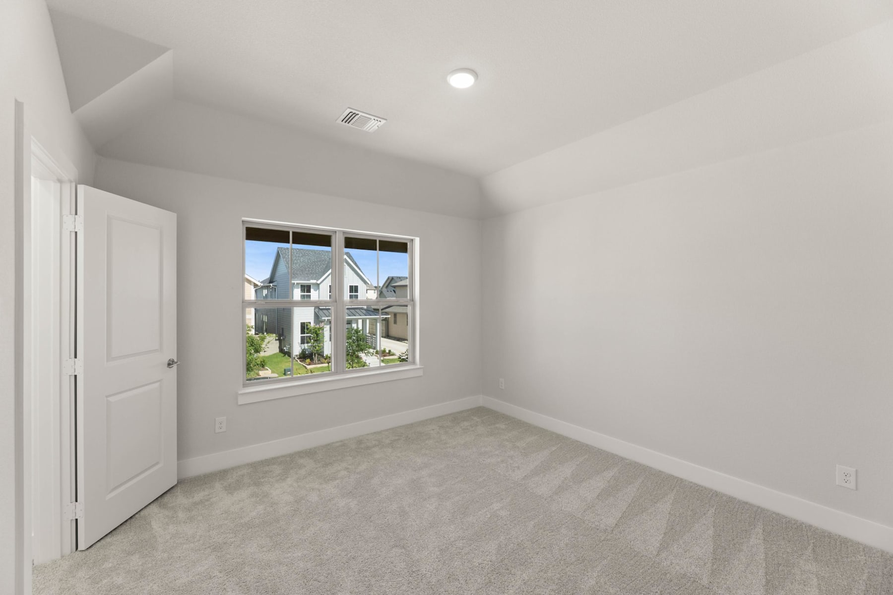 A bright and spacious room with a large window overlooking a residential neighborhood, featuring white walls, a carpeted floor, and a closed door.