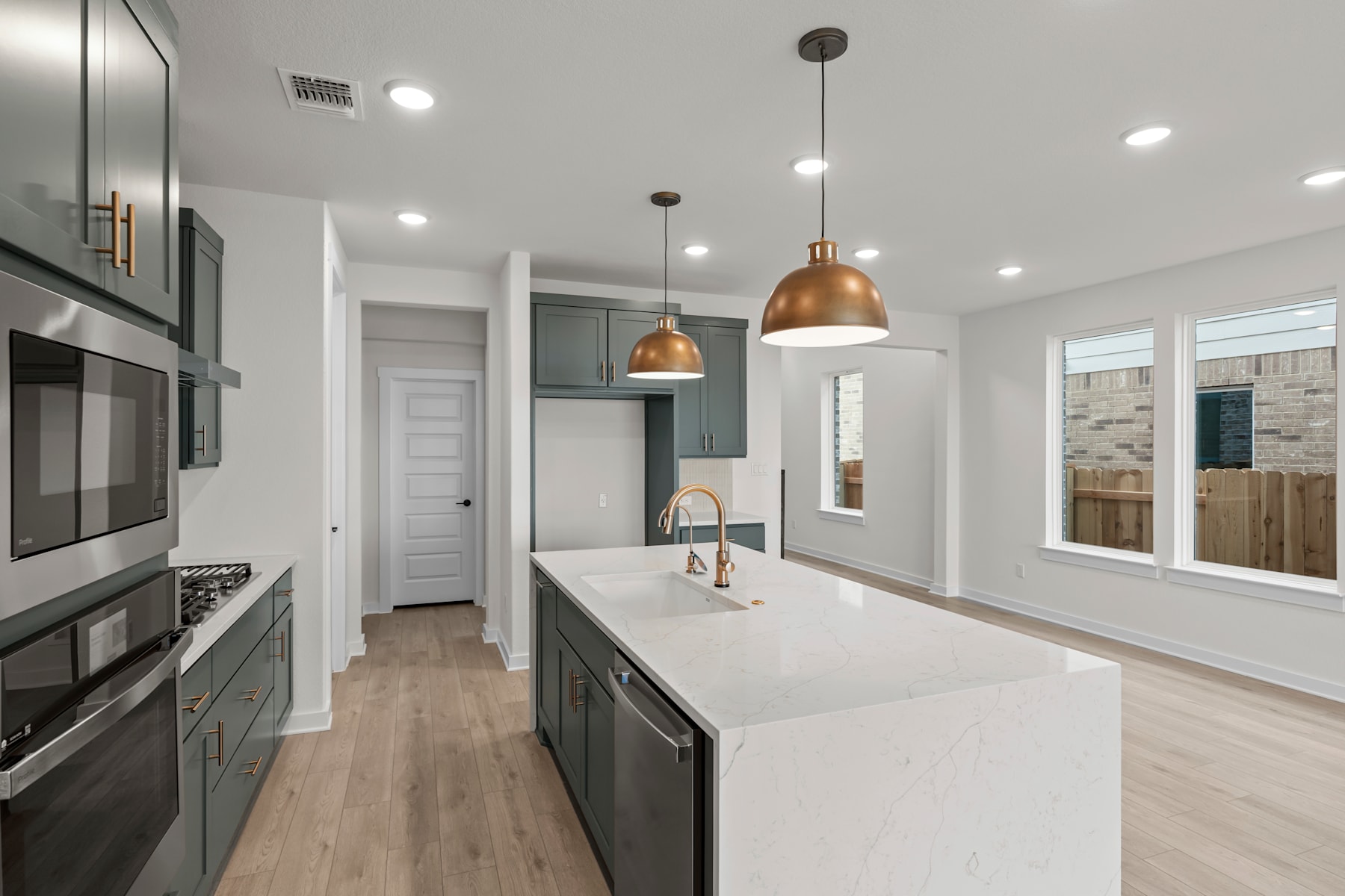 A modern, open-concept kitchen with sleek gray cabinets, white countertops, and copper pendant lights, set against a backdrop of light-colored walls and hardwood floors.