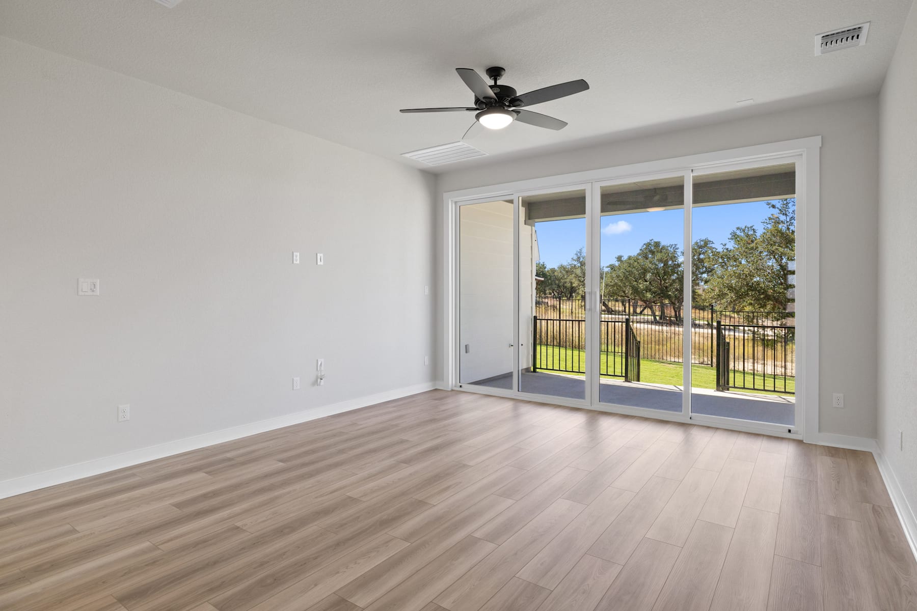 A spacious room with a ceiling fan, large windows overlooking a grassy outdoor area, and hardwood flooring.