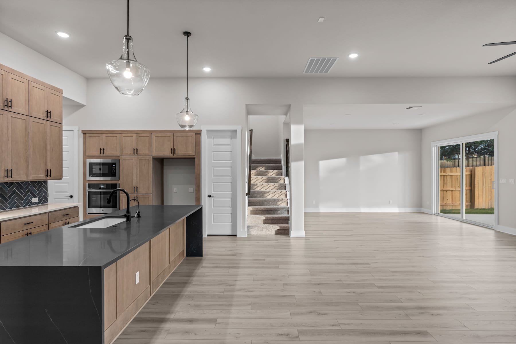 A modern, open-concept kitchen and living space with light-colored wood cabinets, black countertops, and a sliding glass door leading to an outdoor area.
