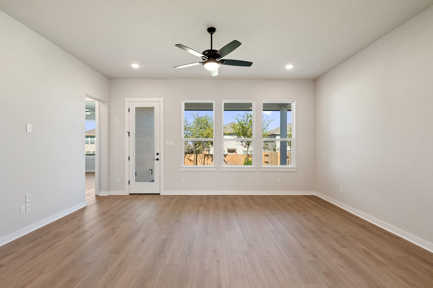 A spacious, well-lit room with a ceiling fan, hardwood floors, and large windows overlooking an outdoor area.