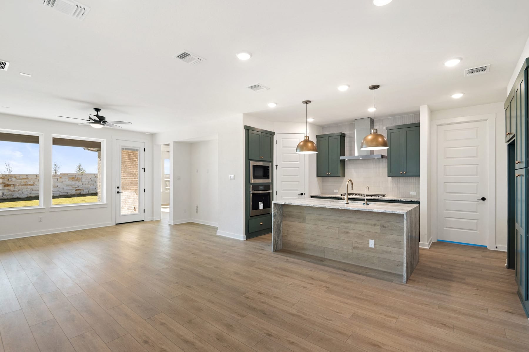 A modern, open-concept kitchen and living space with hardwood floors, white walls, and gray cabinetry, featuring large windows that provide natural light and a view of the outdoors.