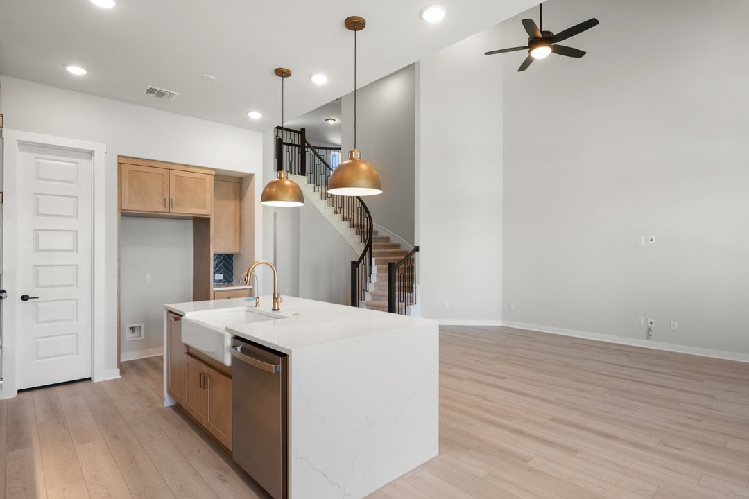 A modern, open-concept kitchen with white countertops, wooden cabinets, and pendant lighting, leading into a living area with a staircase and ceiling fan.