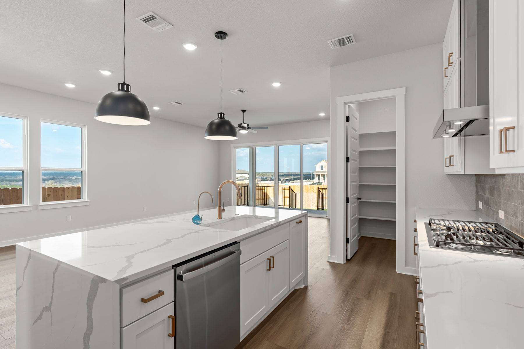 A modern, well-lit kitchen with white cabinets, a marble countertop, and pendant lights, leading to a hallway with shelving and a view of the outdoors.