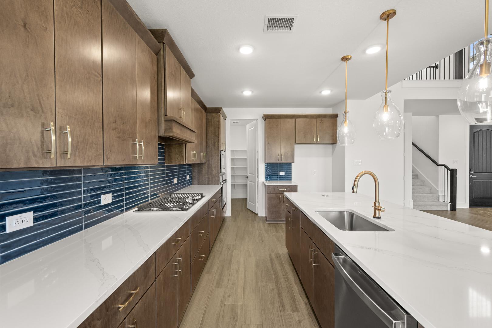 A modern and spacious kitchen with wooden cabinets, white countertops, and a blue tile backsplash, featuring a sink, appliances, and pendant lighting.