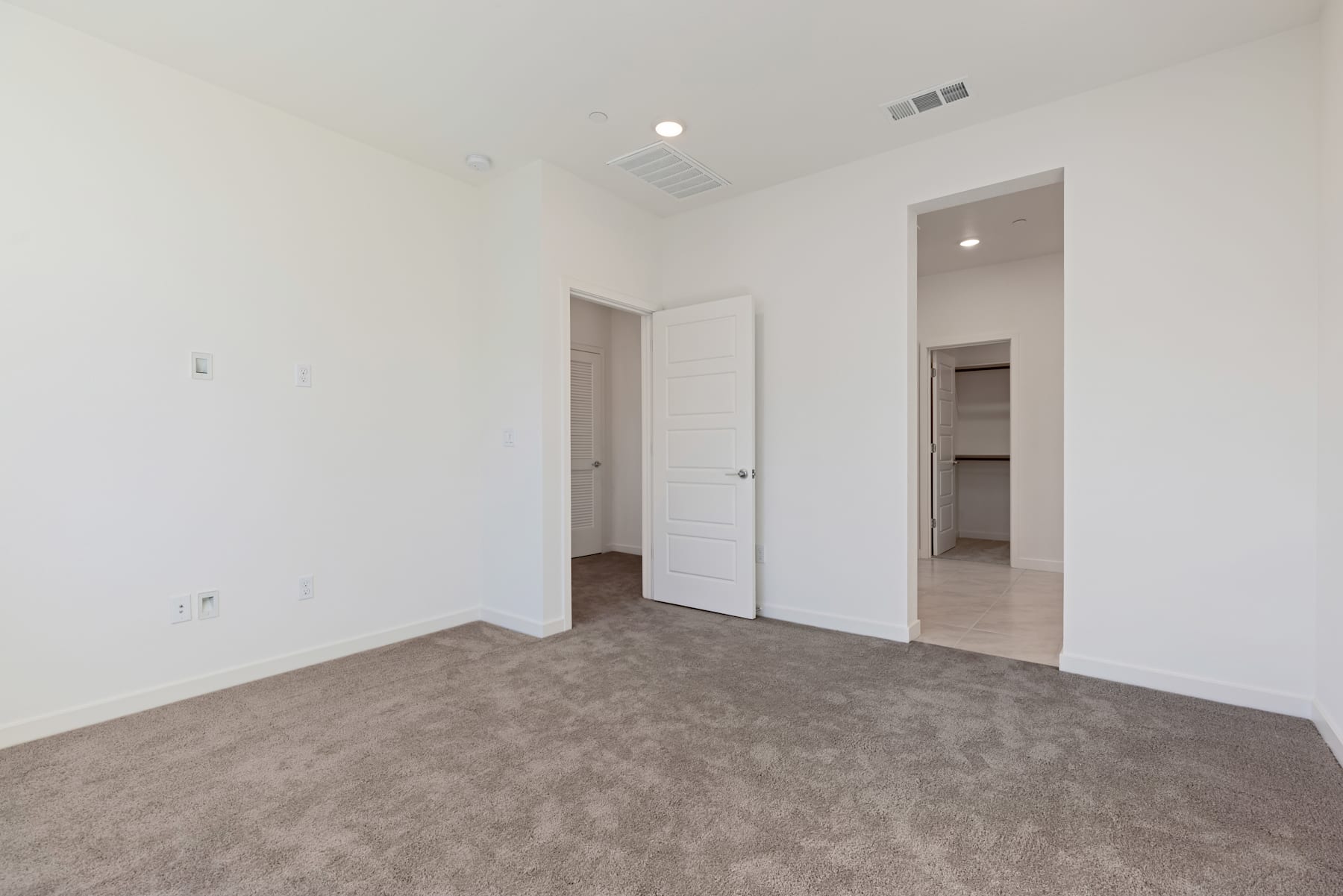 A spacious, minimalist room with white walls, a closed door, and a carpeted floor.