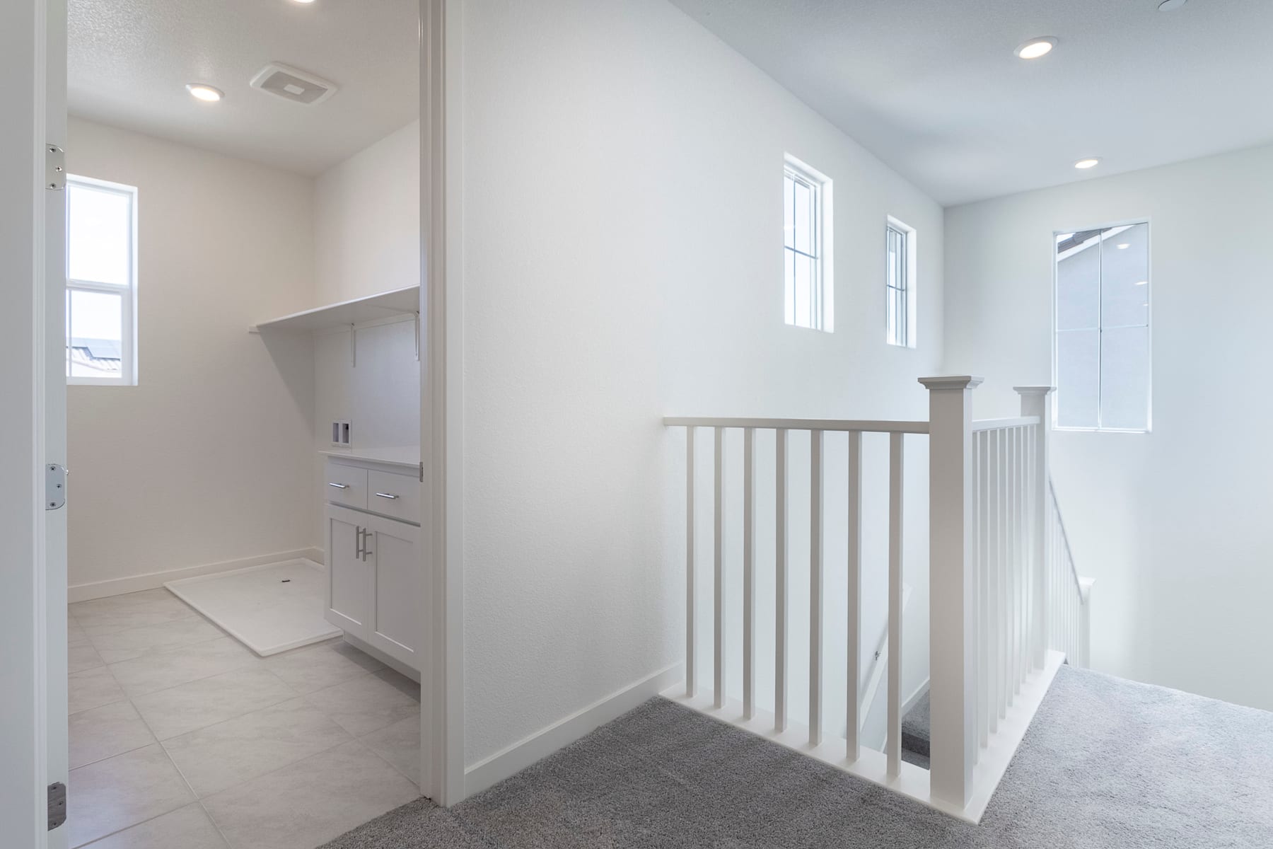 A bright, airy hallway with a white staircase railing leading up to a second floor, and a doorway visible in the foreground.