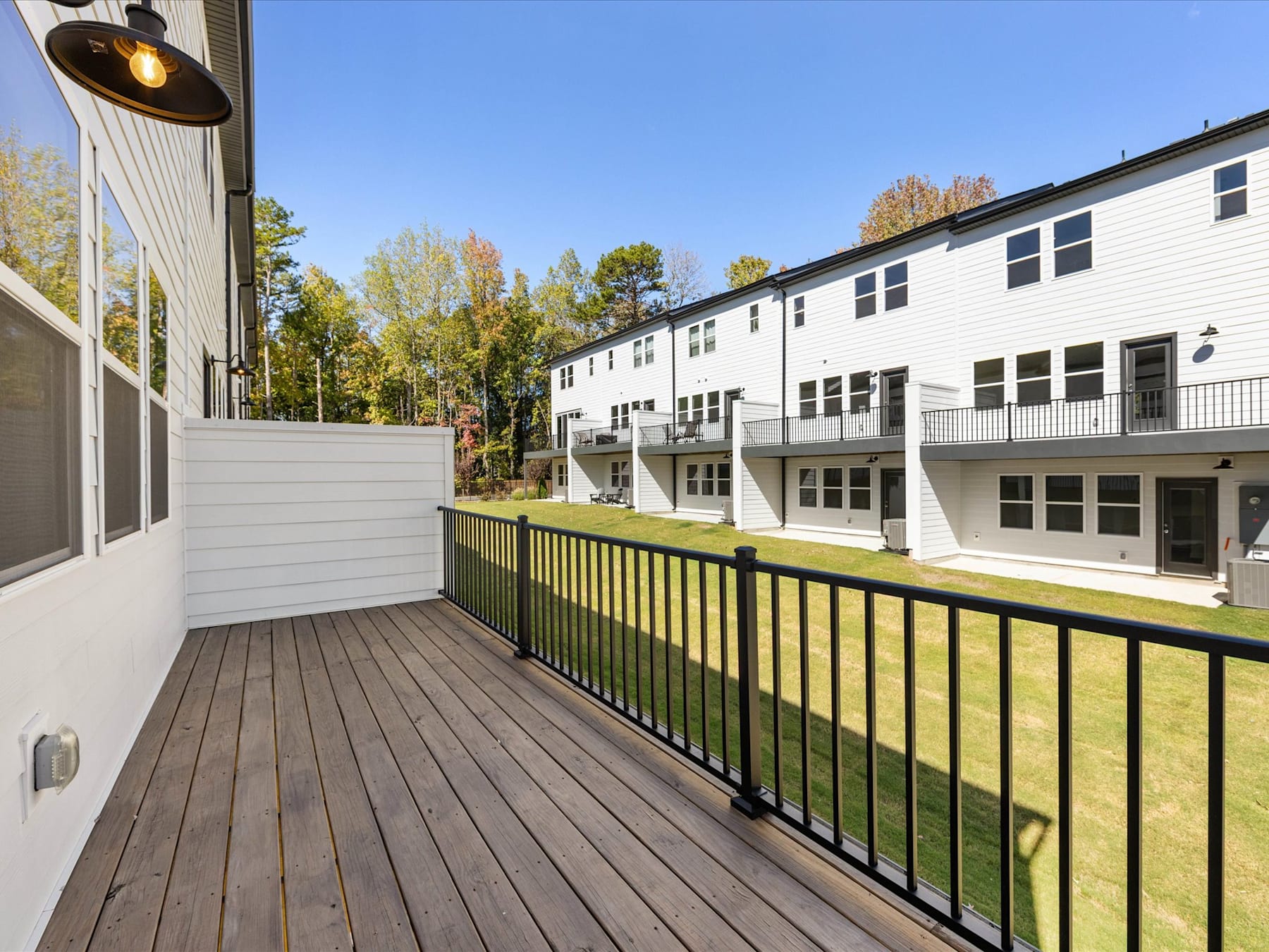 A wooden deck with a railing overlooks a grassy area, surrounded by a row of white apartment buildings with trees in the background.