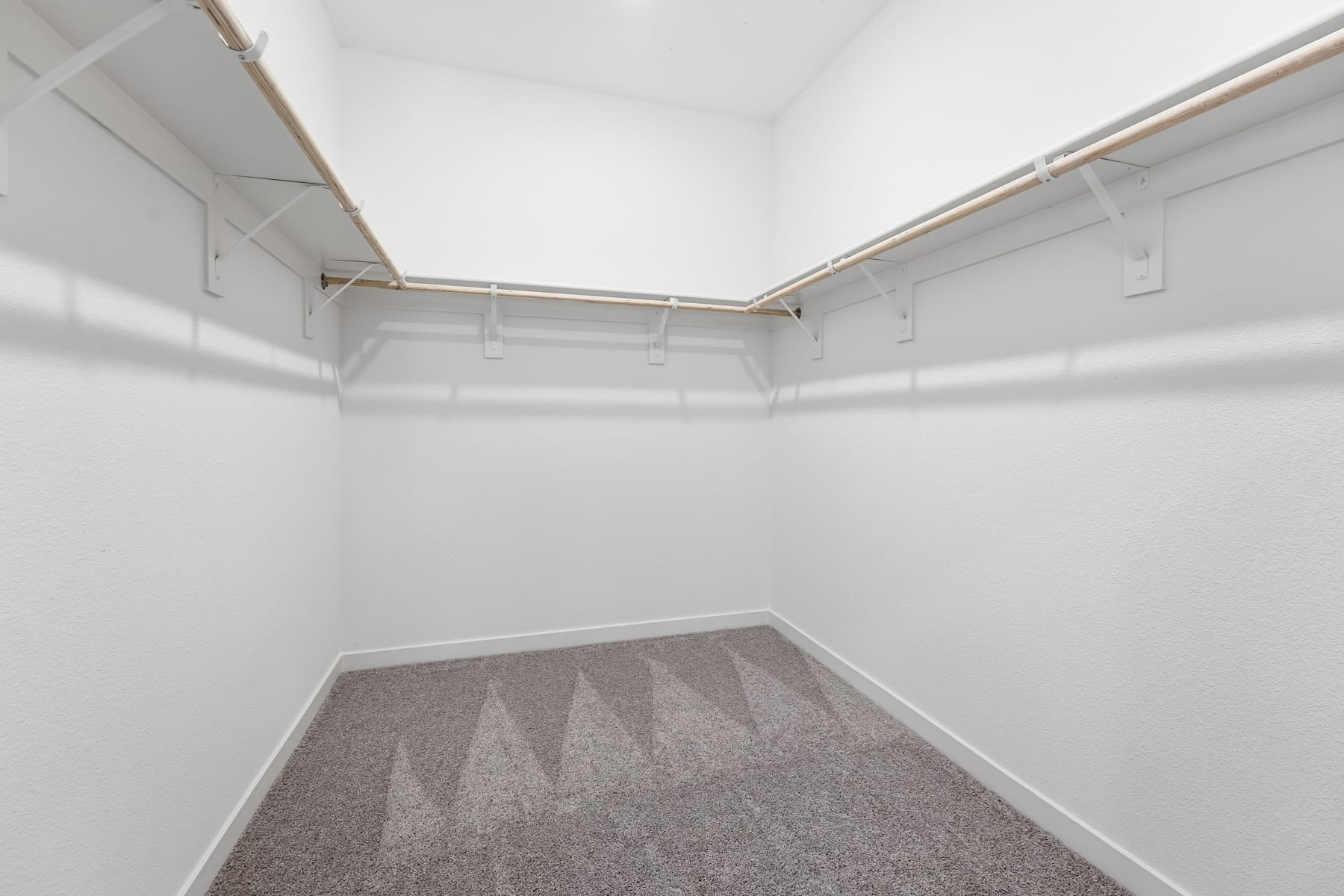 A small, empty room with white walls and a carpeted floor, featuring a metal clothing rod and shelves along the walls.