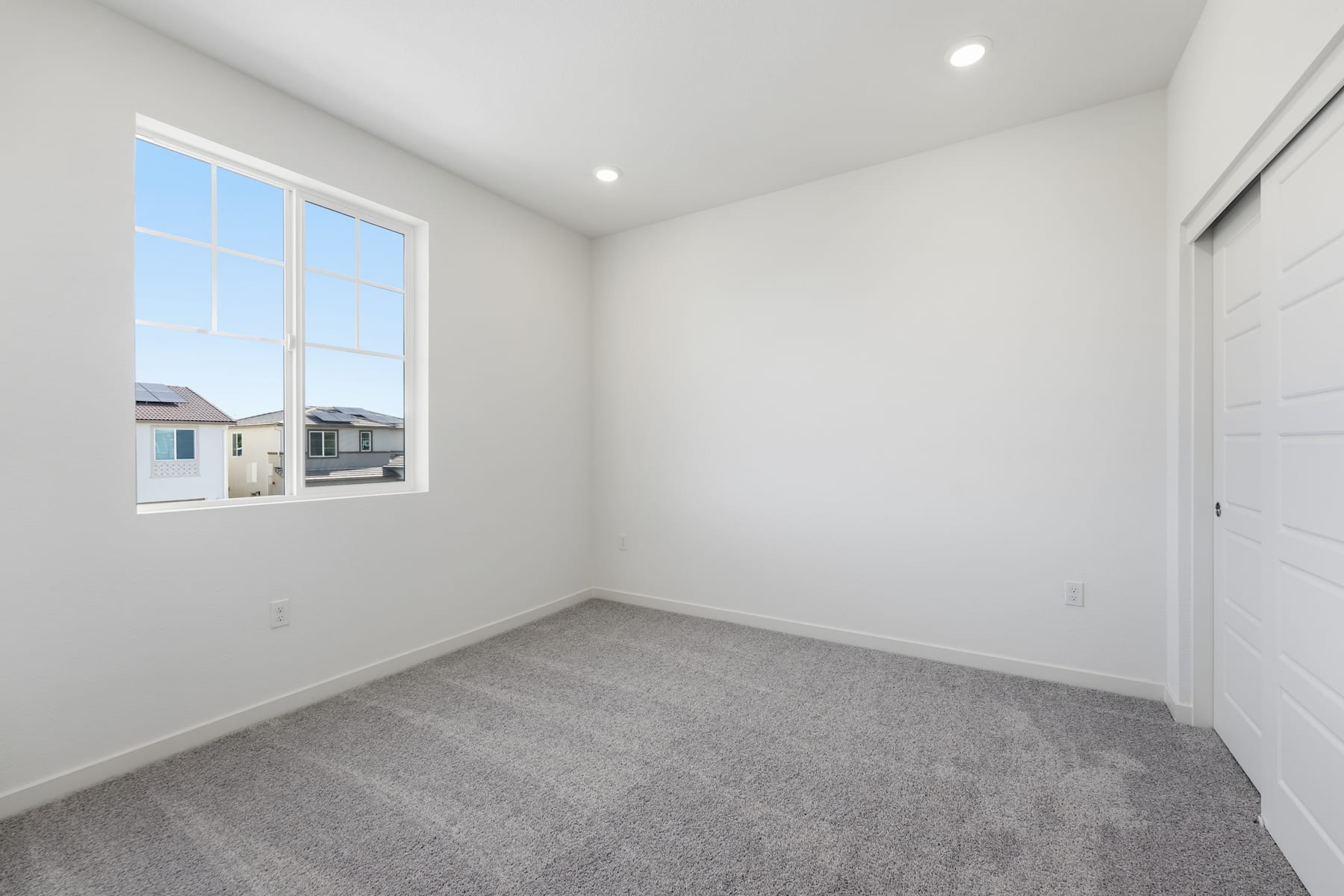 A bright, empty room with a large window overlooking residential buildings in the background, and a gray carpeted floor in the foreground.