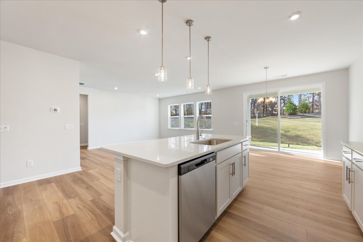 A modern, open-concept kitchen with white cabinets, stainless steel appliances, and a large window overlooking a grassy outdoor area.