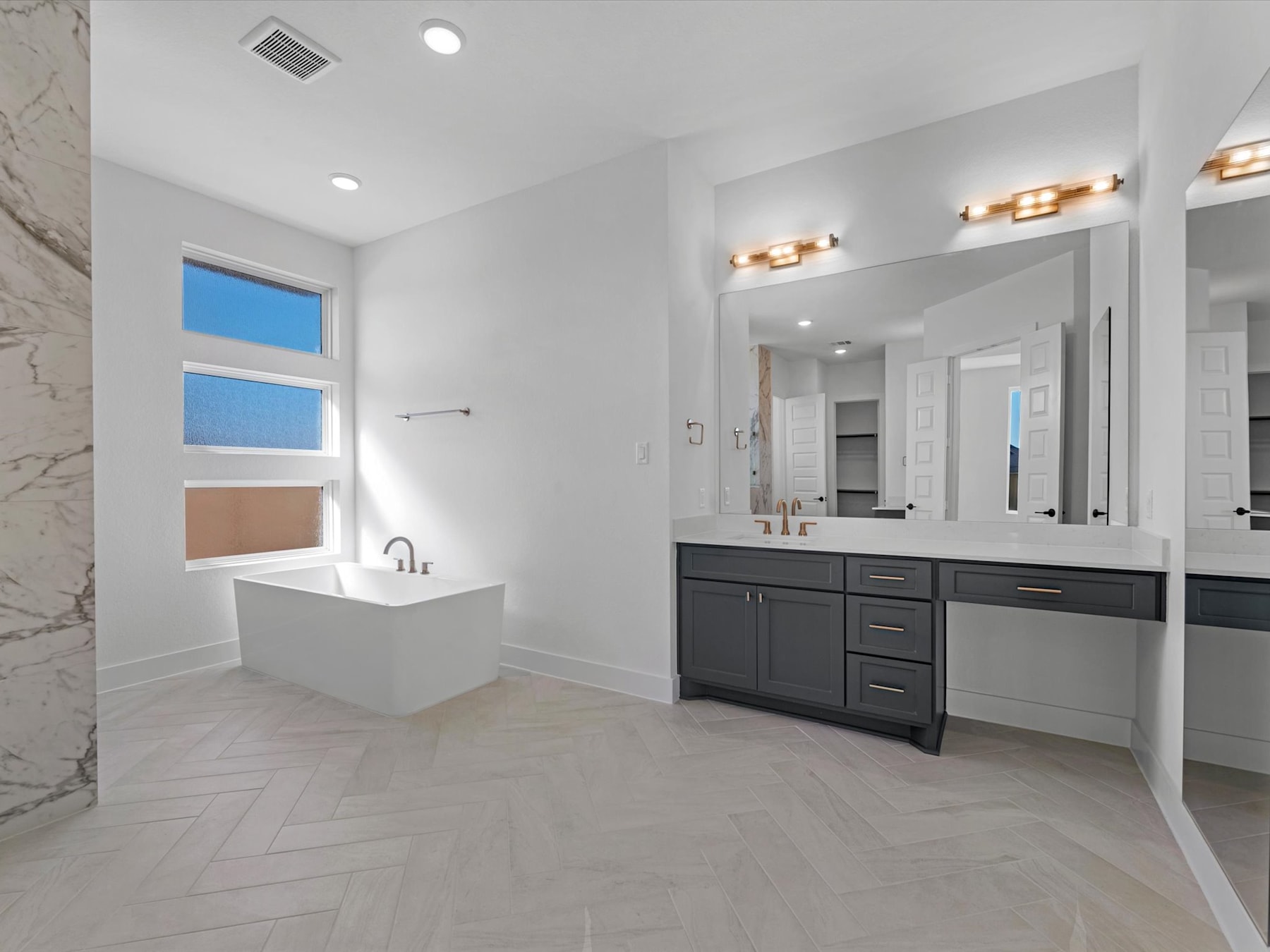 A modern and spacious bathroom with a white freestanding bathtub, a dark vanity cabinet with a large mirror, and a tiled floor in a herringbone pattern.