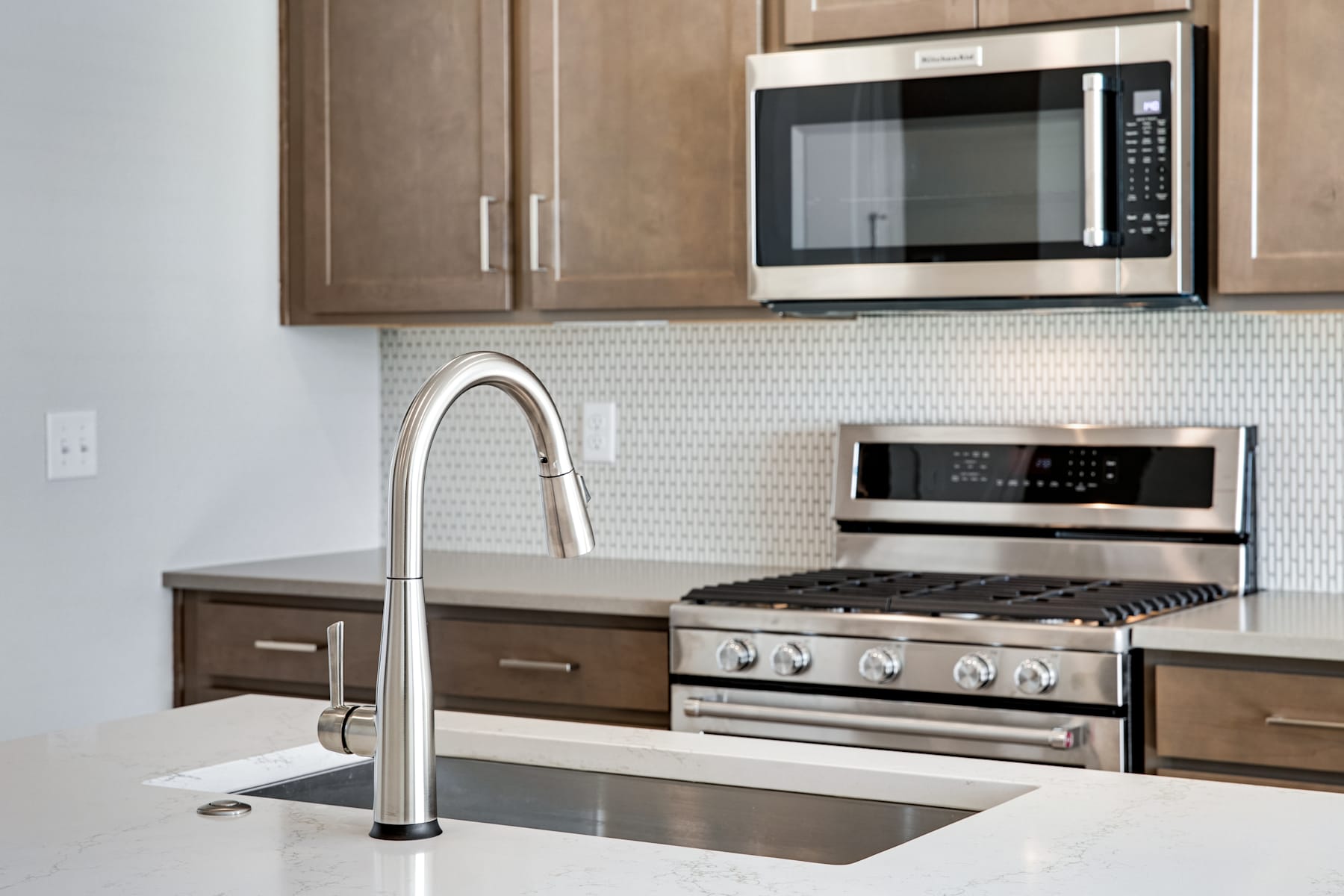 A modern kitchen with wooden cabinets, a stainless steel gas stove, and a sleek faucet in the foreground, along with a microwave oven mounted above the stove in the background.
