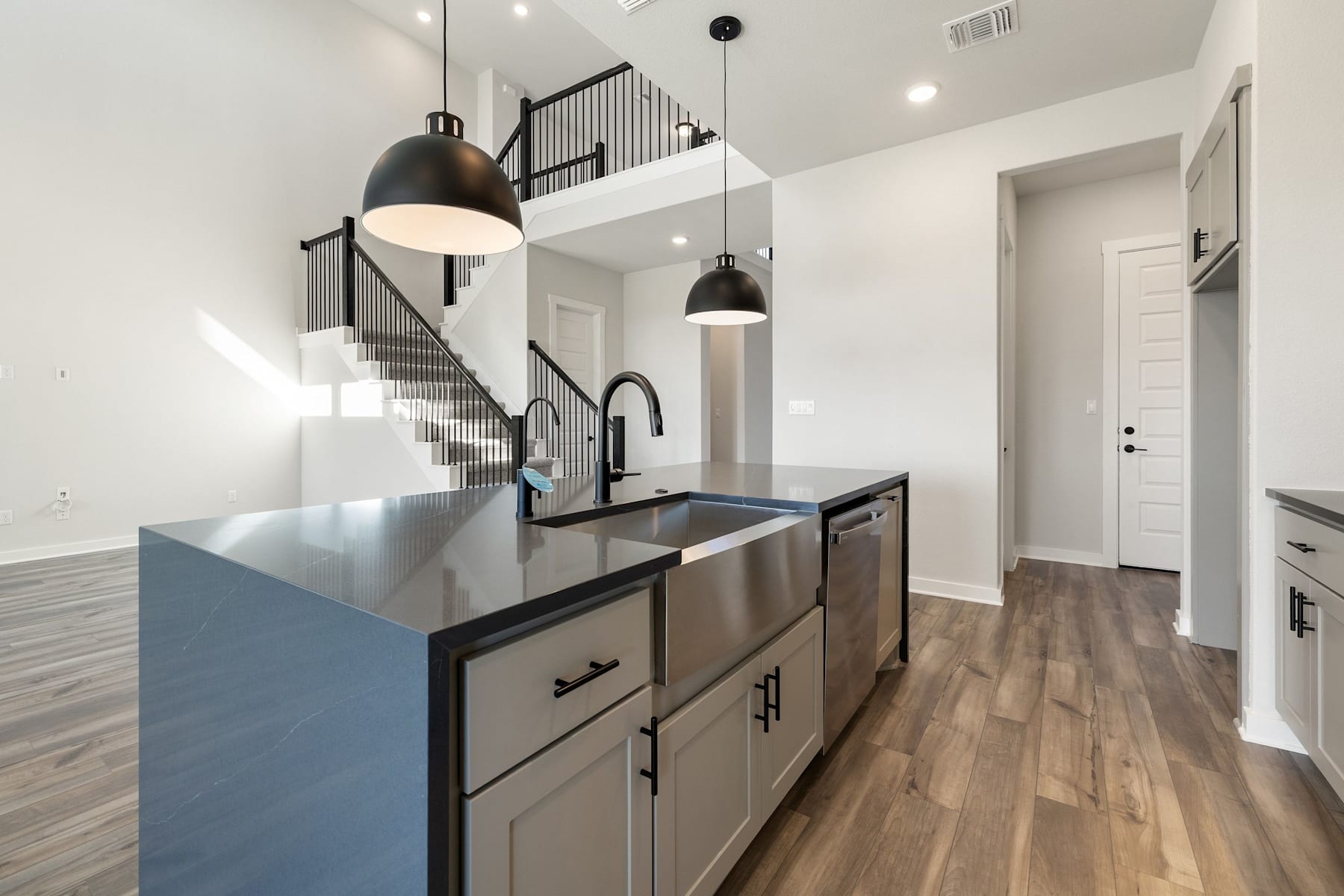 A modern and minimalist kitchen with stainless steel appliances, dark cabinets, and pendant lighting, leading into a hallway with a staircase visible in the background.