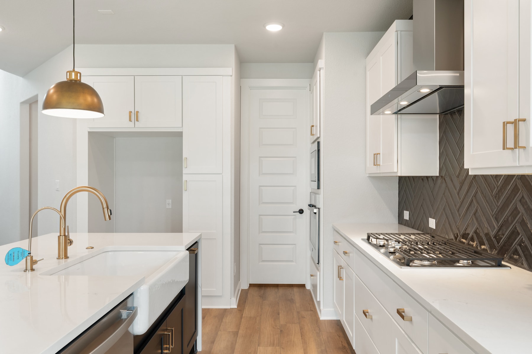 A modern, well-lit kitchen with white cabinets, a gold pendant light, and a herringbone tile backsplash, featuring a sink and stove in the foreground.