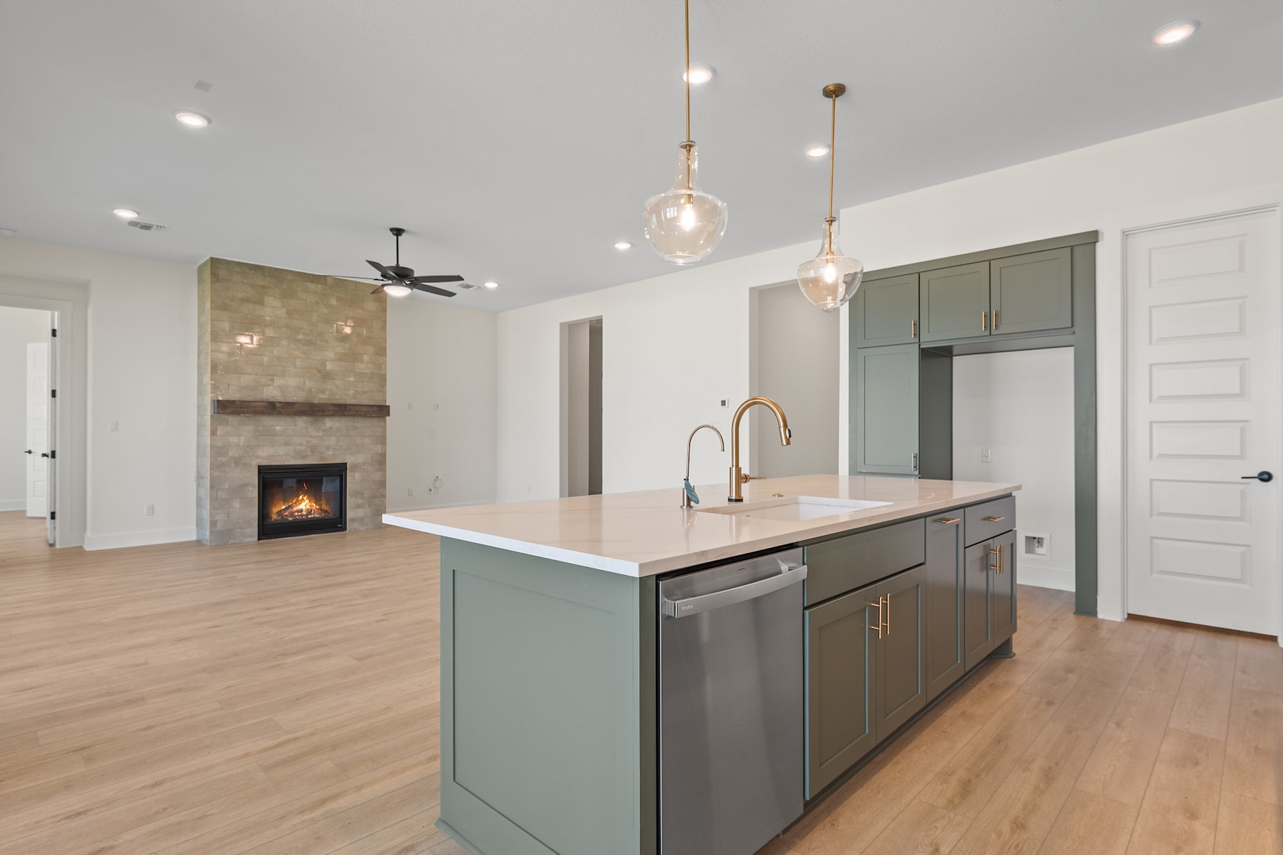 A modern and spacious kitchen with a central island, hardwood floors, and a fireplace in the background, illuminated by pendant lights.