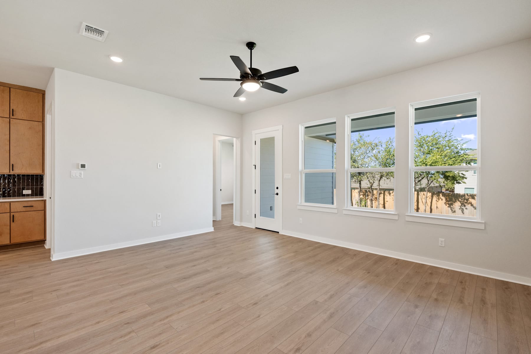 A spacious, well-lit room with a ceiling fan, wooden floors, and large windows overlooking a lush outdoor environment.