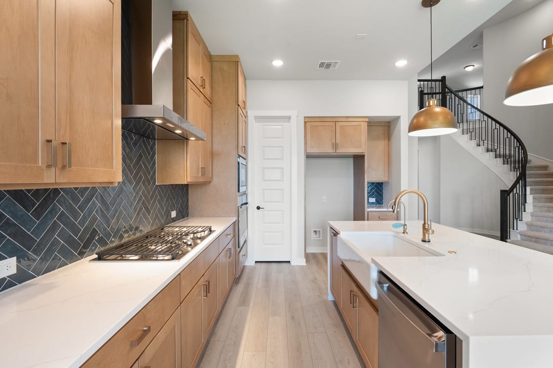 A modern and well-designed kitchen with light wood cabinets, a white countertop, and a herringbone tile backsplash, leading into a hallway with a staircase.