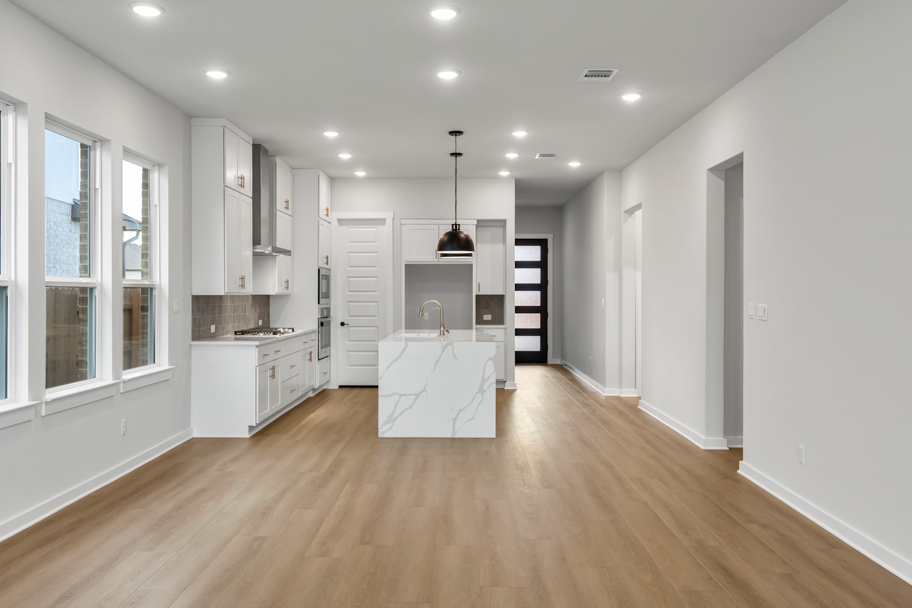 A modern, bright, and spacious kitchen with white cabinets, a large marble-topped island, and hardwood floors extending into the adjacent hallway.