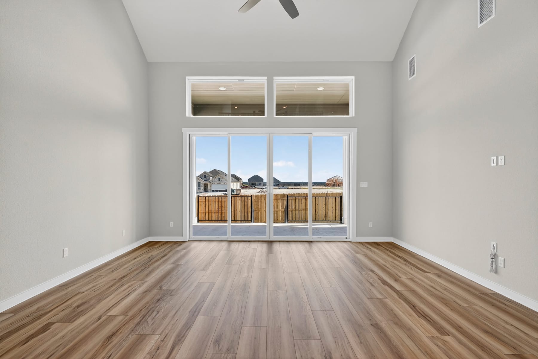 A spacious room with large windows overlooking a backyard, featuring hardwood floors and a high ceiling with a ceiling fan.