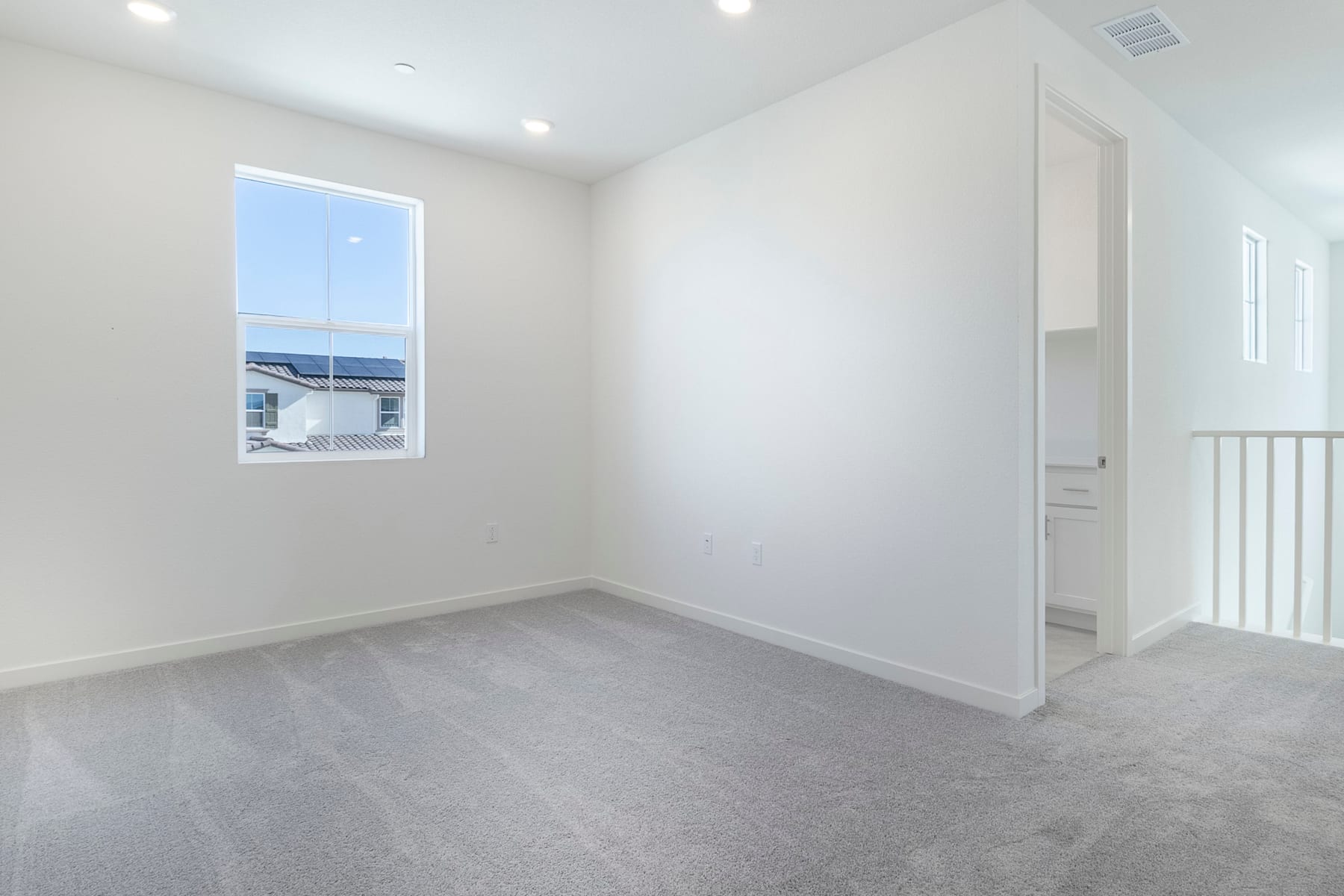 A bright, empty room with white walls, a window, and a gray carpeted floor.