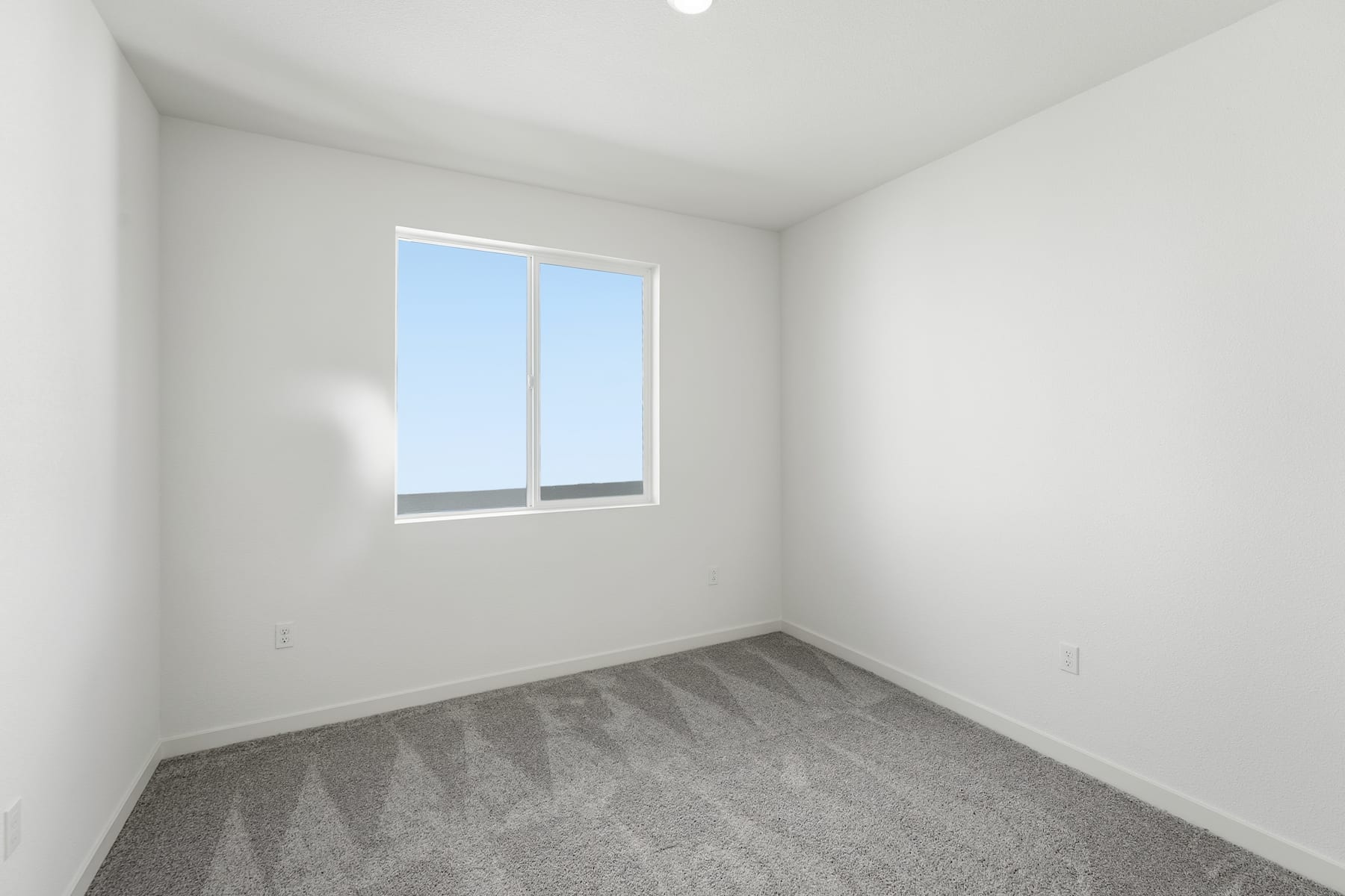 A simple, minimalist room with a window overlooking the sky, featuring a gray carpeted floor and plain white walls.