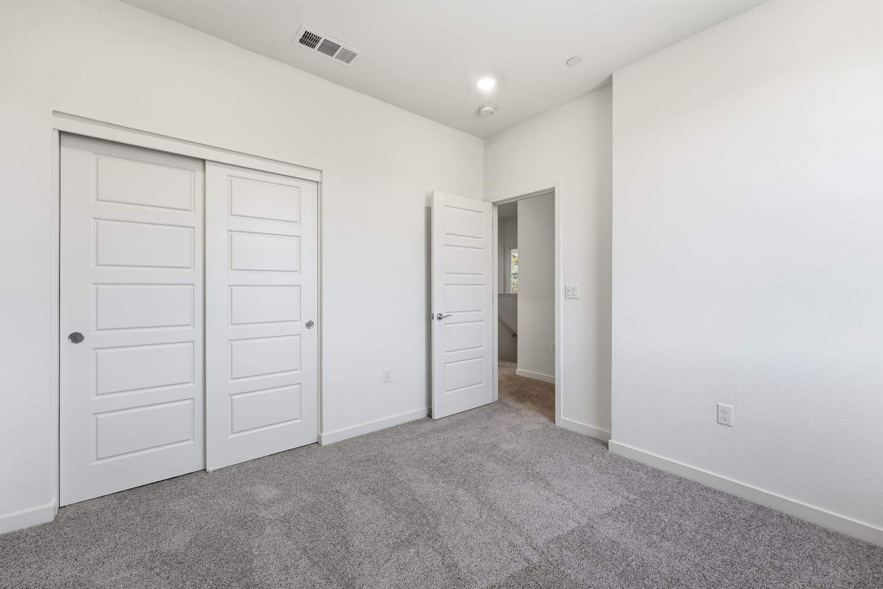 A spacious, minimalist bedroom with white walls, gray carpeting, and two white doors leading to other rooms.